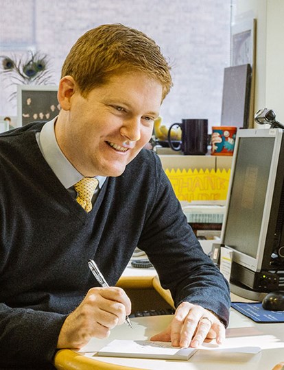 Man in jumper vest sat at a desk chatting to another person and writing things down